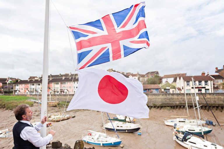 Two flags (Union Jack and Japanese) being raised on a flagpole in Pill, North Somerset by the father of the groom. Boats lay on the mud in the estuary behind and colourful houses on the other side of the estuary. Photograph by Blooming Photography.