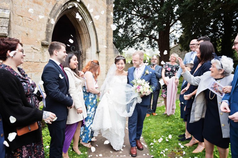 Candid photograph of bride and groom exiting the church whilst being showered with confetti. Story telling photograph by Blooming Photography.