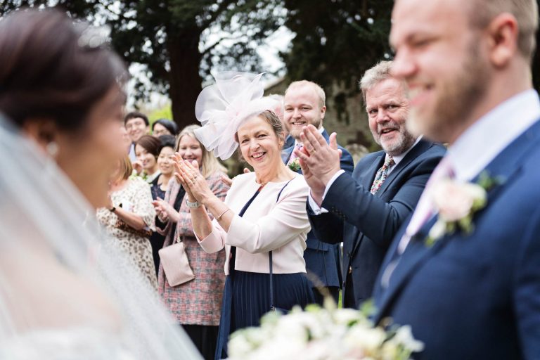 Parents clapping in focus in the background with the bride and groom smiling out of focus at the foreground. Story telling photograph by Blooming Photography.
