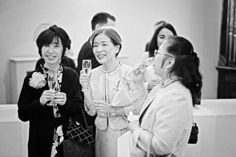 Guests sip champagne, chat and look onward at other guests. Candid black and white photograph by Blooming Photography.
