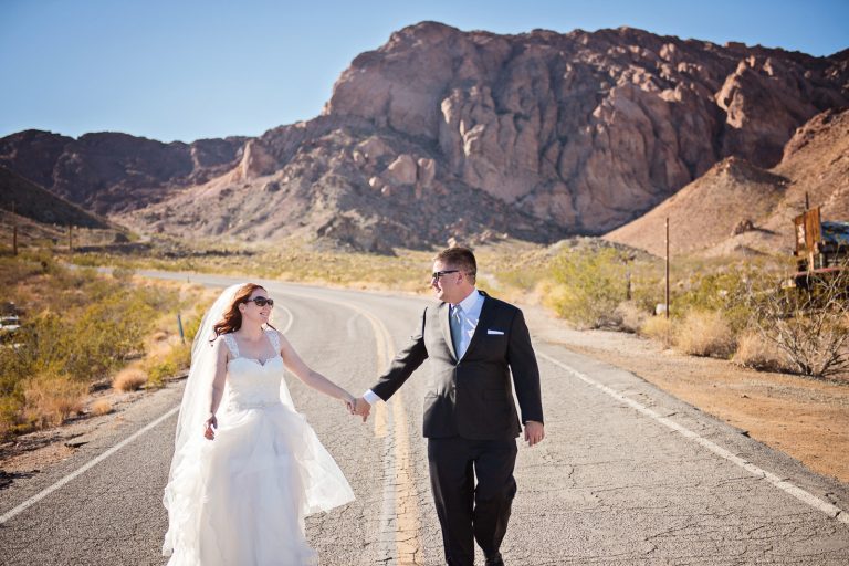 Bride and groom walk and in hand away along the highway in Nevada with rocks behind them Photograph by Blooming Photography, photographed at Nelson Ghost Town.