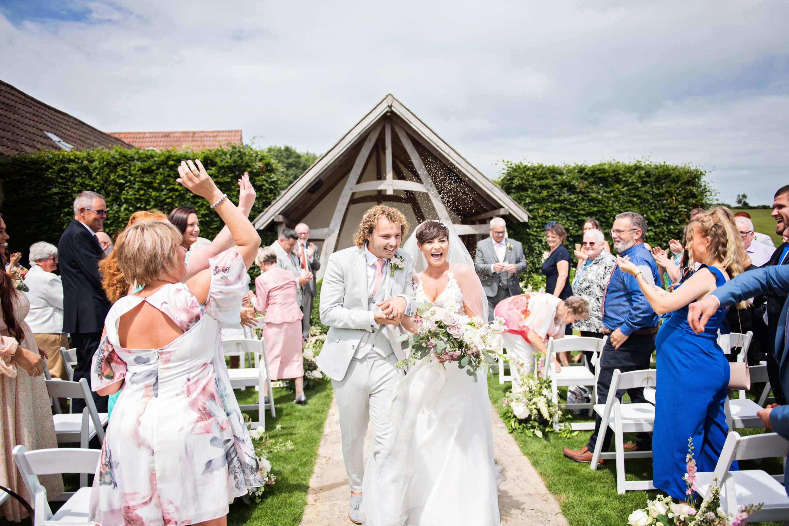 Colourful image of a bride and groom walking down the wedding isle (outside) at Kingscote Barn, whilst being showered with confetti from their wedding guests. Candid photo by wedding photographer Blooming Photography.