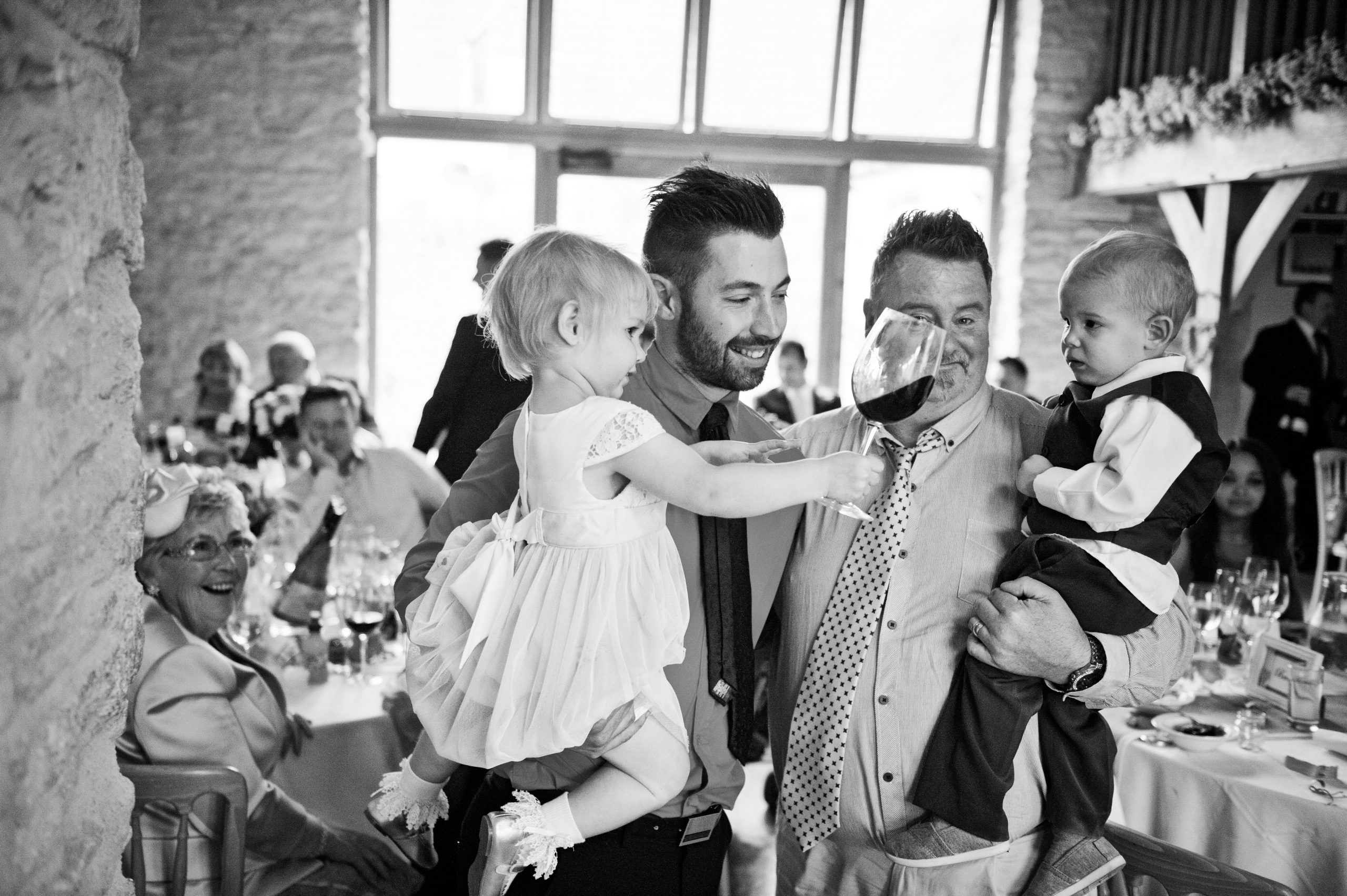 B&W image of two dads at wedding carrying two young children. The youngest child (girl), is holding a large glass of red wine and is offering it to the young boy.Both dads are smiling.
