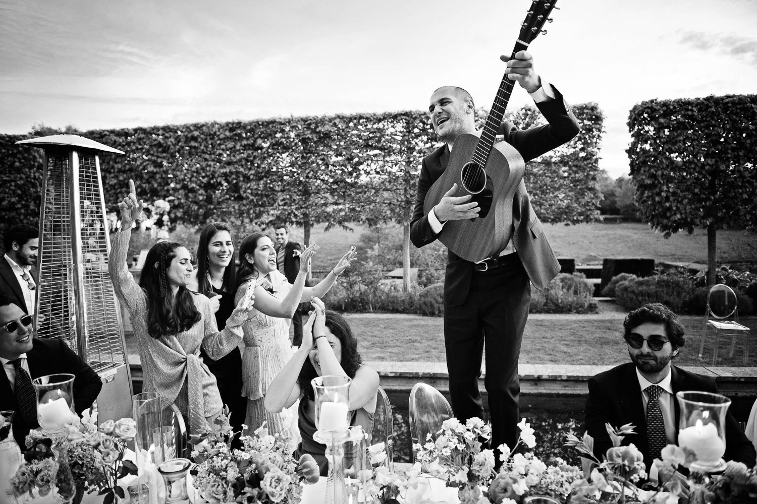 Candid documentary style photo of wedding guest standing on a chair playing his guitar, wedding guests are partying.